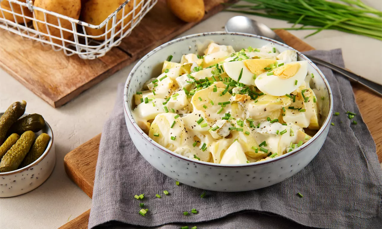 Kartoffelsalat mit cremigem Dressing, garniert mit frischen Schnittlauchröllchen und Eischeiben in einer gesprenkelten Schale auf grauem Tuch