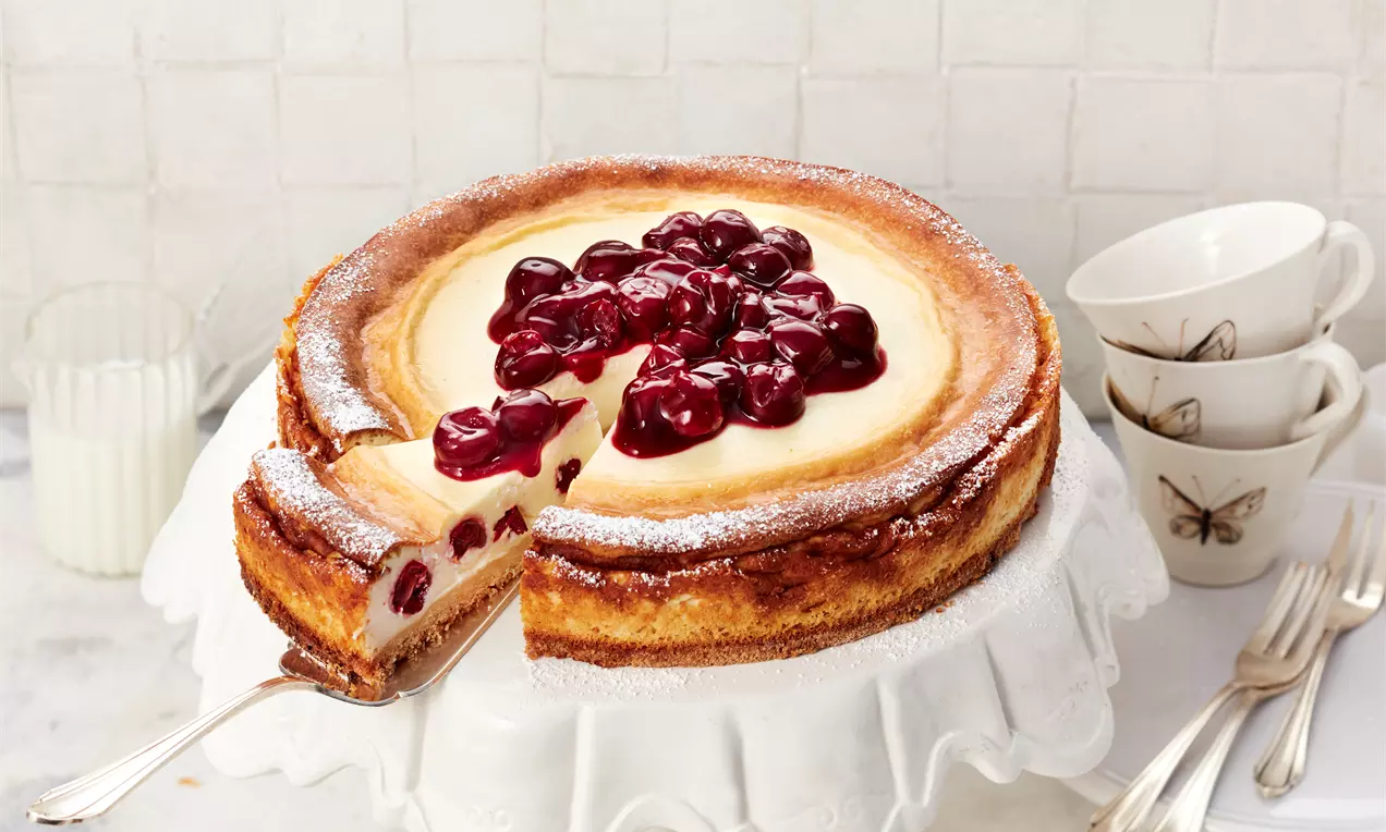 Goldbrauner Käsekuchen mit heller, cremiger Füllung und glänzenden roten Kirschen auf einer weißen Tortenplatte mit herausgehobener Kuchenscheibe