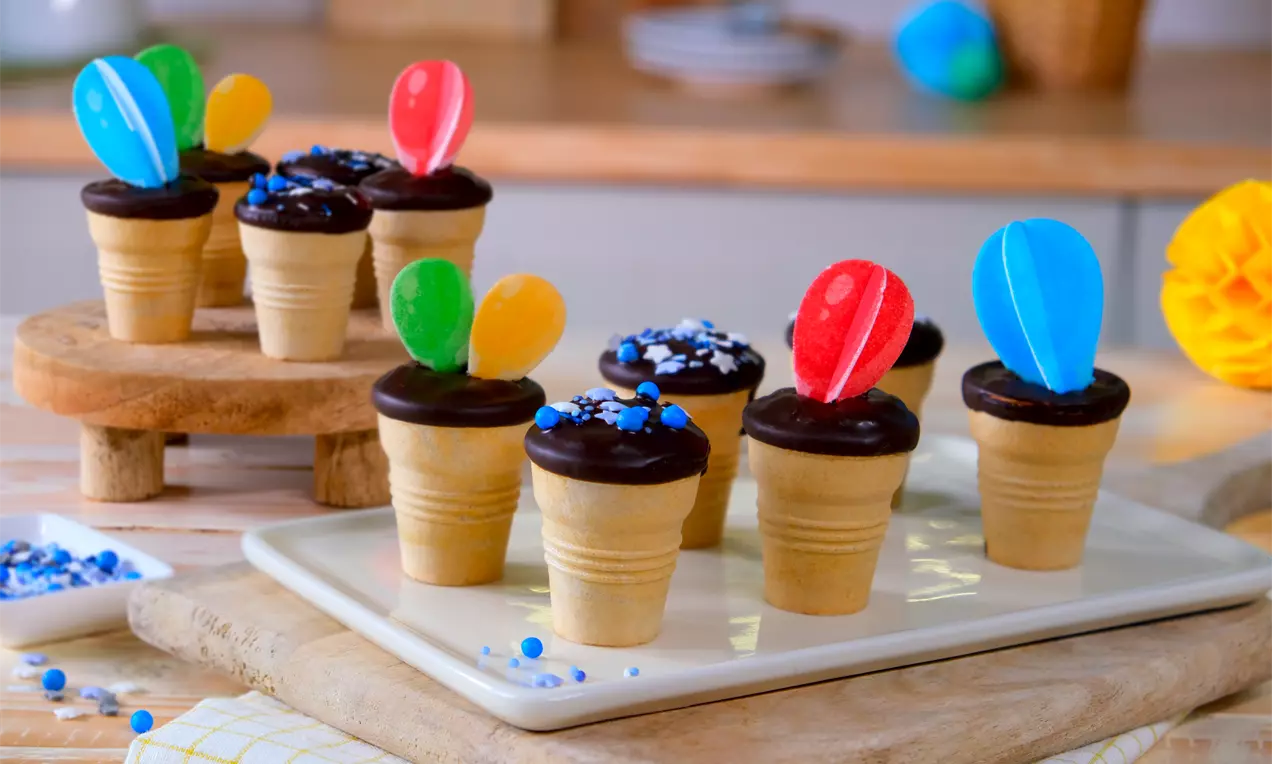 Kleine Waffelbecher mit dunkler Glasur, bunten Ballon-Dekoren und Zuckerstreuseln auf Holzbrett und Teller arrangiert