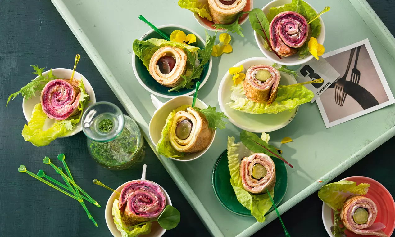 Bunte Pfannkuchen-Röllchen mit farbigen Füllungen, auf Salatblättern serviert, dekorativ auf kleinen Tellern und einem grünen Tablett arrangiert