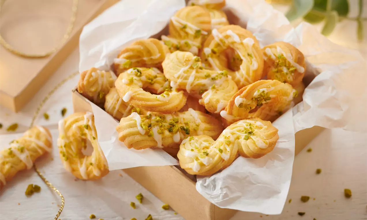 Goldgelbe spritzförmige Orangenplätzchen mit weißem Zuckerguss und gehackten Pistazien, angerichtet in einer Schachtel mit Backpapier