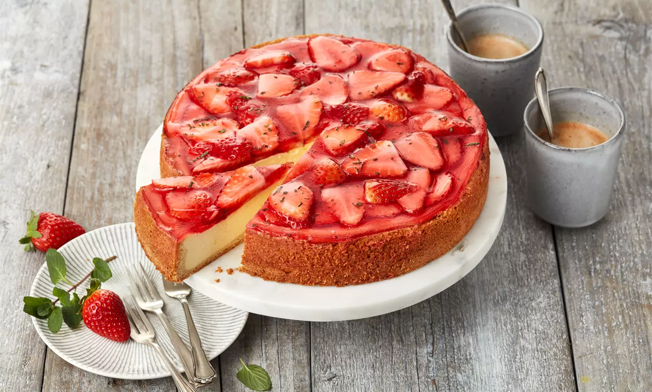 Ein runder Käsekuchen mit goldbraunem Boden, glänzend rotem Erdbeerspiegel und frischen Erdbeeren auf weißer Platte mit Besteck und Tassen