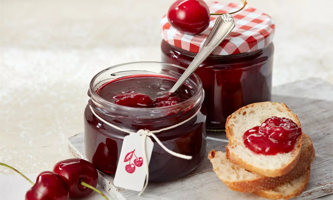 Tiefrote Sauerkirschmarmelade in einem offenen Glas mit silberner Löffelspitze, daneben ein Glas mit rot-weiß kariertem Deckel und Kirschen