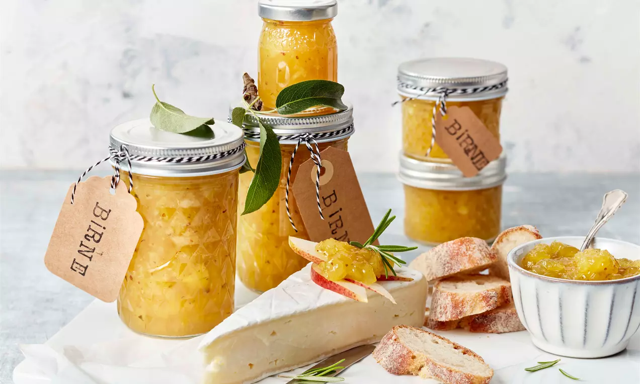 Goldgelbes Birnenchutney in Gläsern mit Etiketten, drapiert neben Brie, Brotscheiben und einer Schale Chutney auf weißem Untergrund