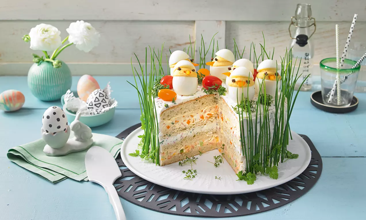 Pikante Schichttorte mit Cremefüllung, dekoriert mit Schnittlauch, Tomaten und weißen Eierküken auf einer weißen Platte mit zartem Grün
