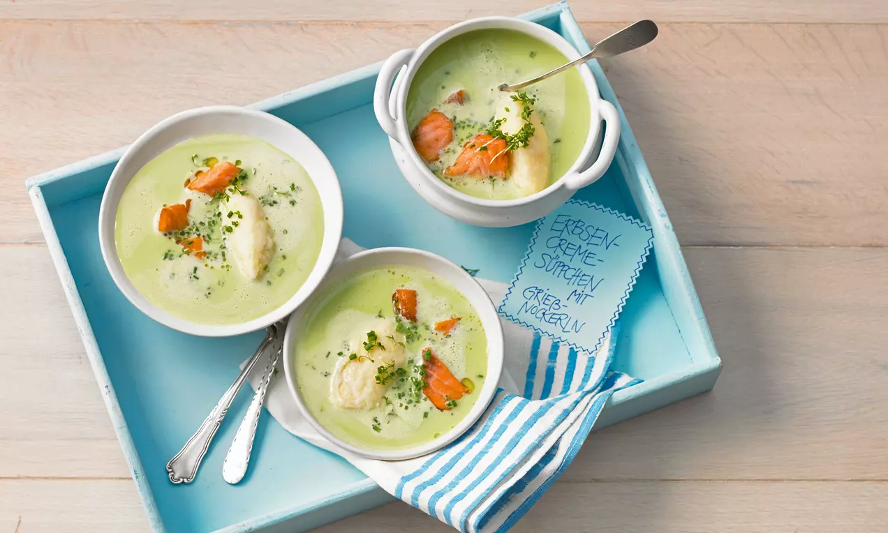 Zartgrüne Erbsencremesuppe mit fluffigen Grießnockerln, orangefarbenen Karottenstückchen und Kräutern in weißen Schälchen angerichtet