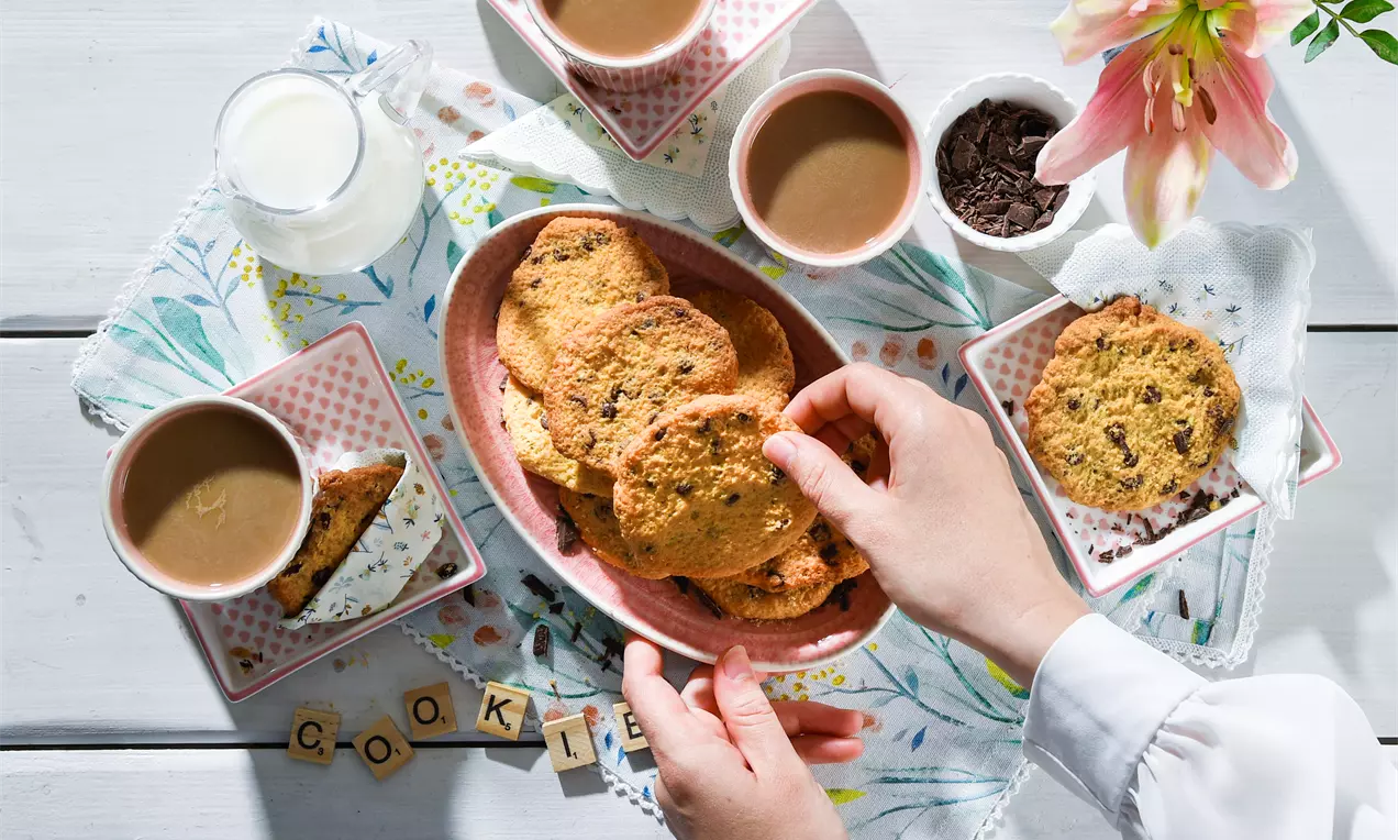 Goldbraune, runde Low-Carb-Plätzchen auf rosafarbener Schale, umgeben von Tassen Kaffee und einer weißen Blume auf hellem Holztisch