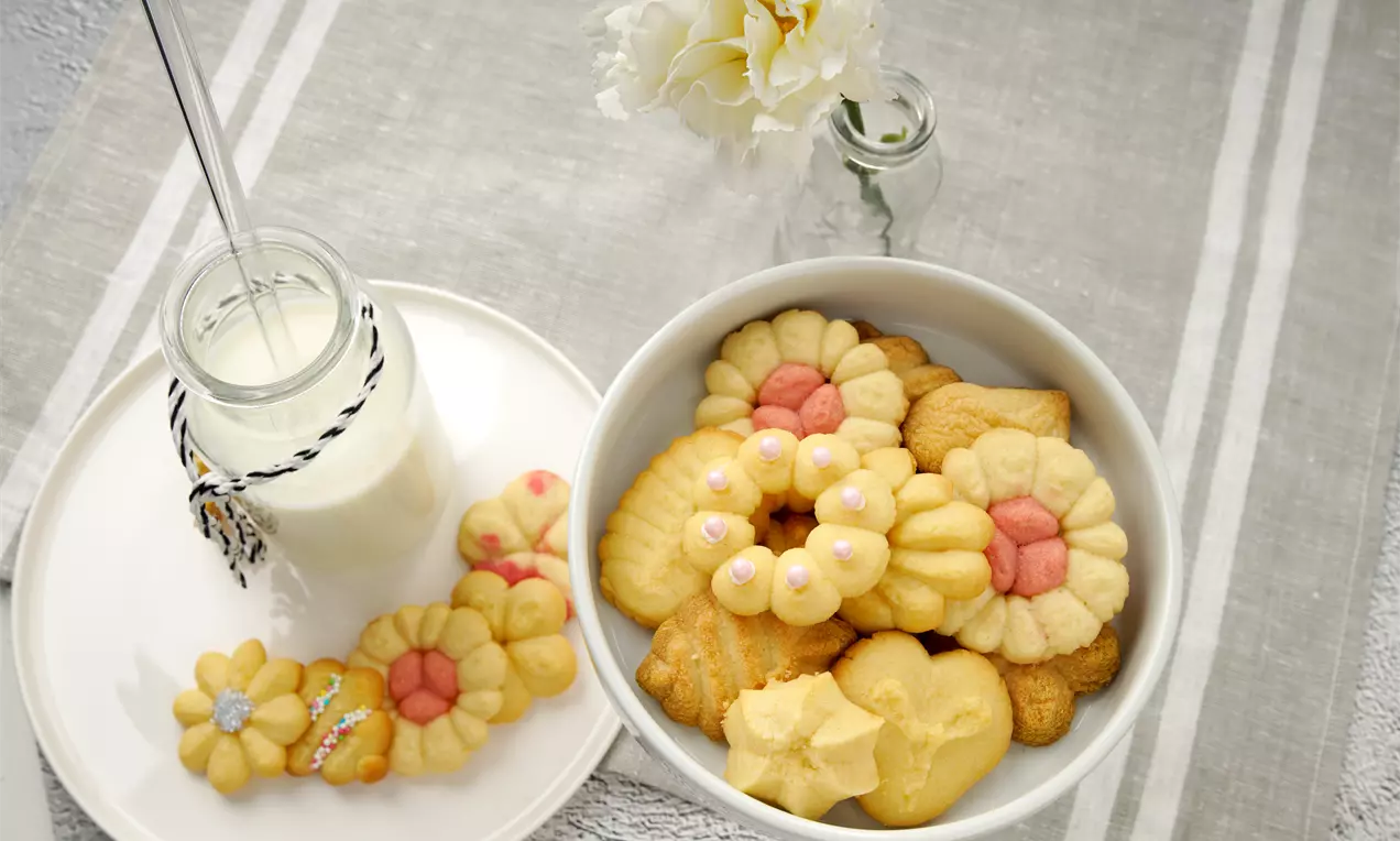 Goldgelbe Eierlikörplätzchen aus der Gebäckpresse in Blumen- und Herzform mit rosa Akzenten in einer weißen Schüssel neben Milchflasche und Blüte