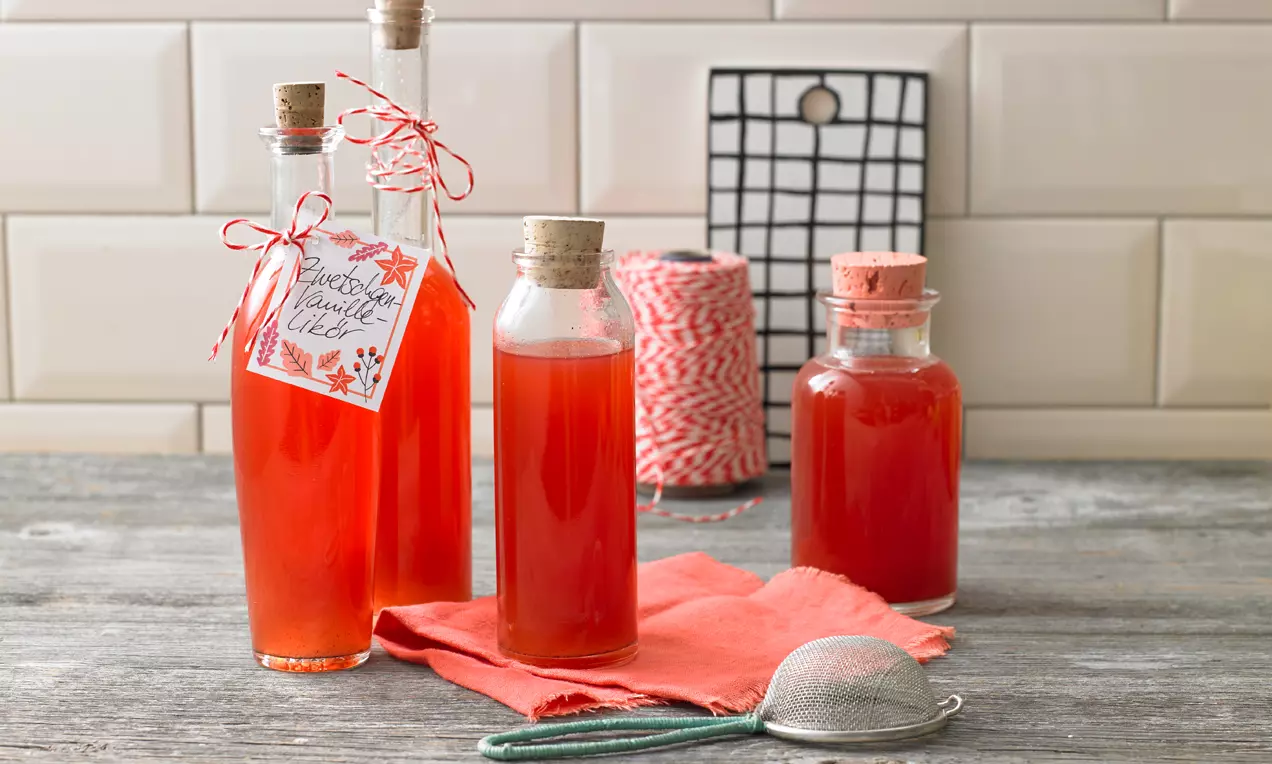 Klare Glasflaschen mit kräftig rotem Zwetschgen-Vanille-Likör, verziert mit Korken und Bändern, arrangiert auf grauem Holztisch mit Küchenutensilien