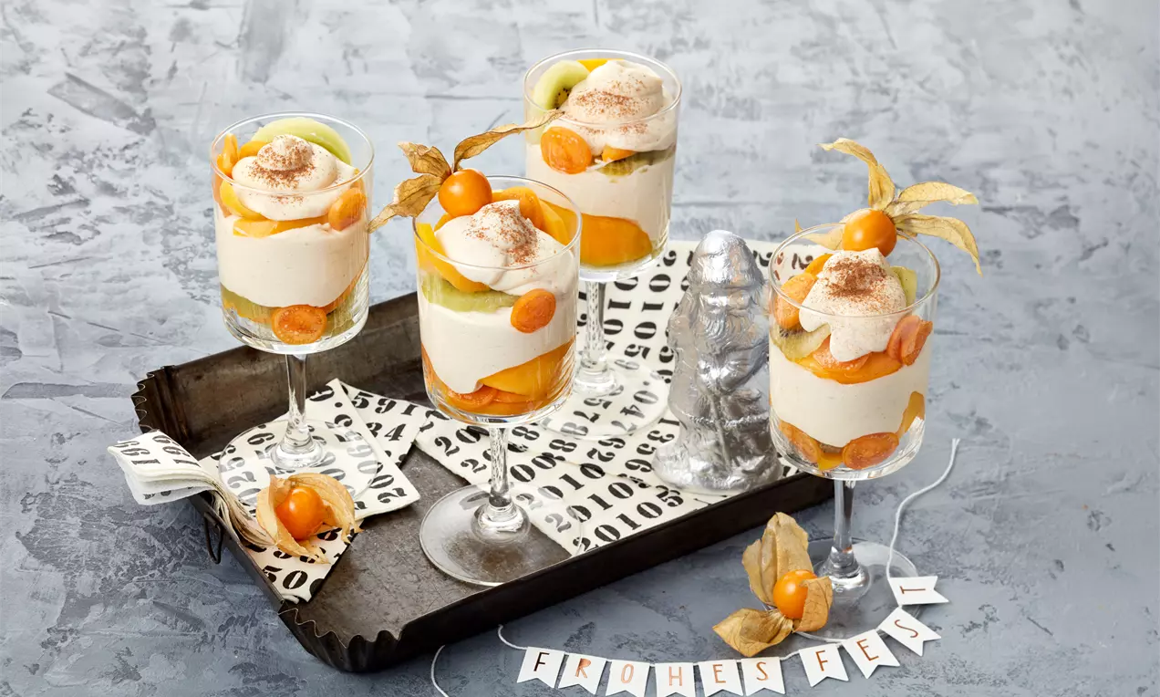 Gläser mit geschichteter Zimt-Orangencreme, gelben Physalis und grünem Obst, dekoriert auf einem Tablett mit festlichem Papier drapiert