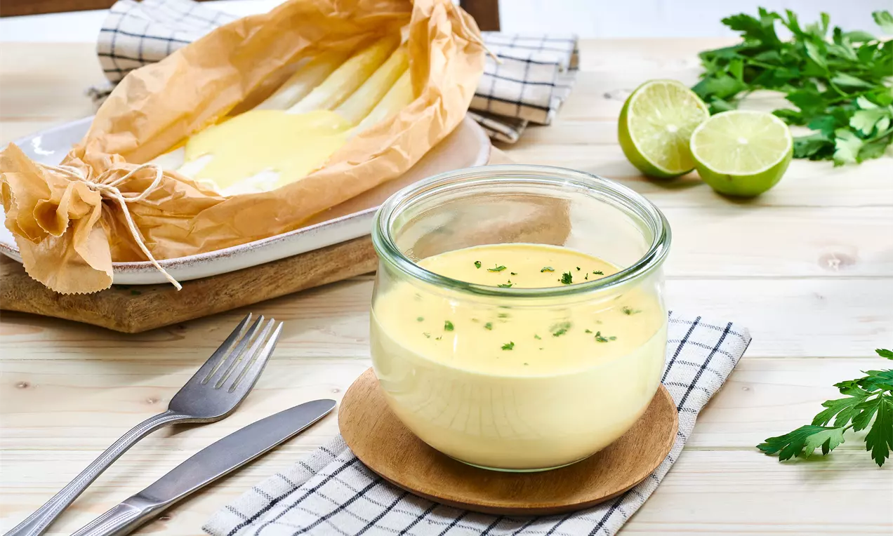 Hellgelbe, cremige Soße mit feinen grünen Kräutern in einem Glas auf einem Holzuntersetzer neben Spargel in Backpapier und Limetten