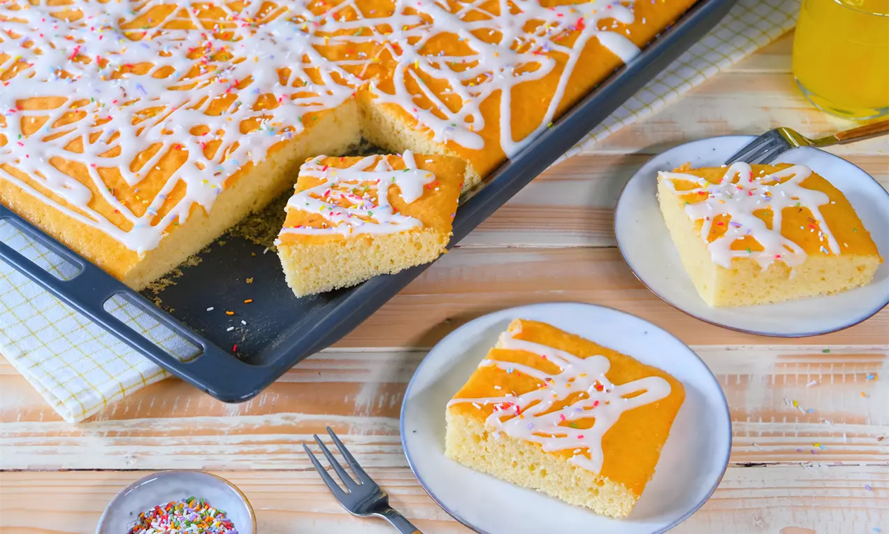 Saftiger, gelboranger Blechkuchen mit weißer Zuckerglasur und bunten Streuseln, angerichtet auf Tellern neben einer Gabel und einem Glas