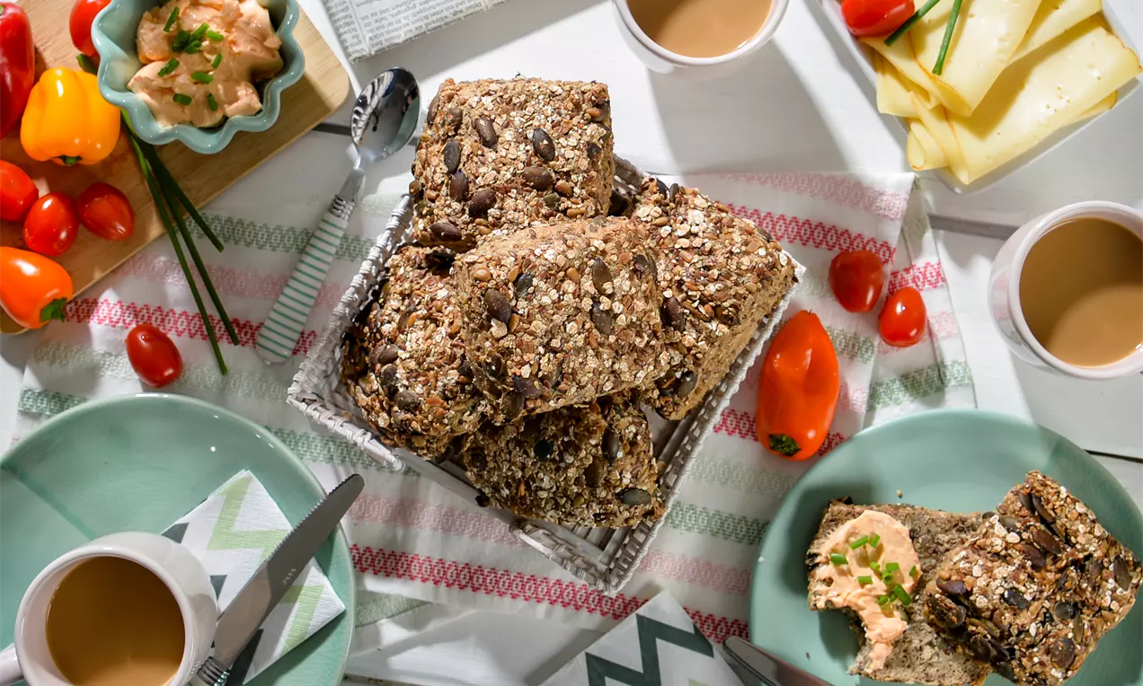 Körnige Mehrkornbrötchen mit goldbrauner Kruste und Saaten, gestapelt in einem kleinen Korb, umgeben von Gemüse, Käse und Kaffee