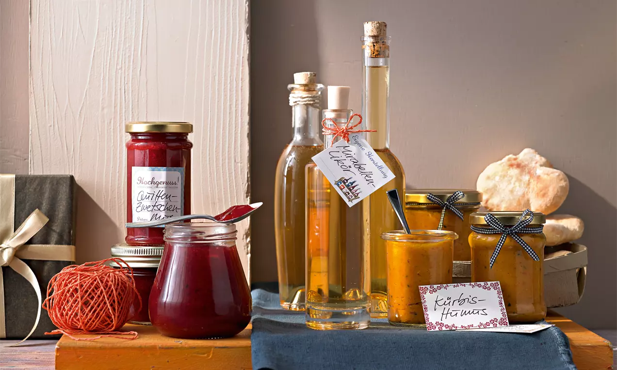 Quitten-Zwetschgen-Mus in Glas mit rotem Deckel neben Mirabellen-Likör in schlanken Flaschen und Kürbis-Hummus in Gläsern mit Stoffdeckel