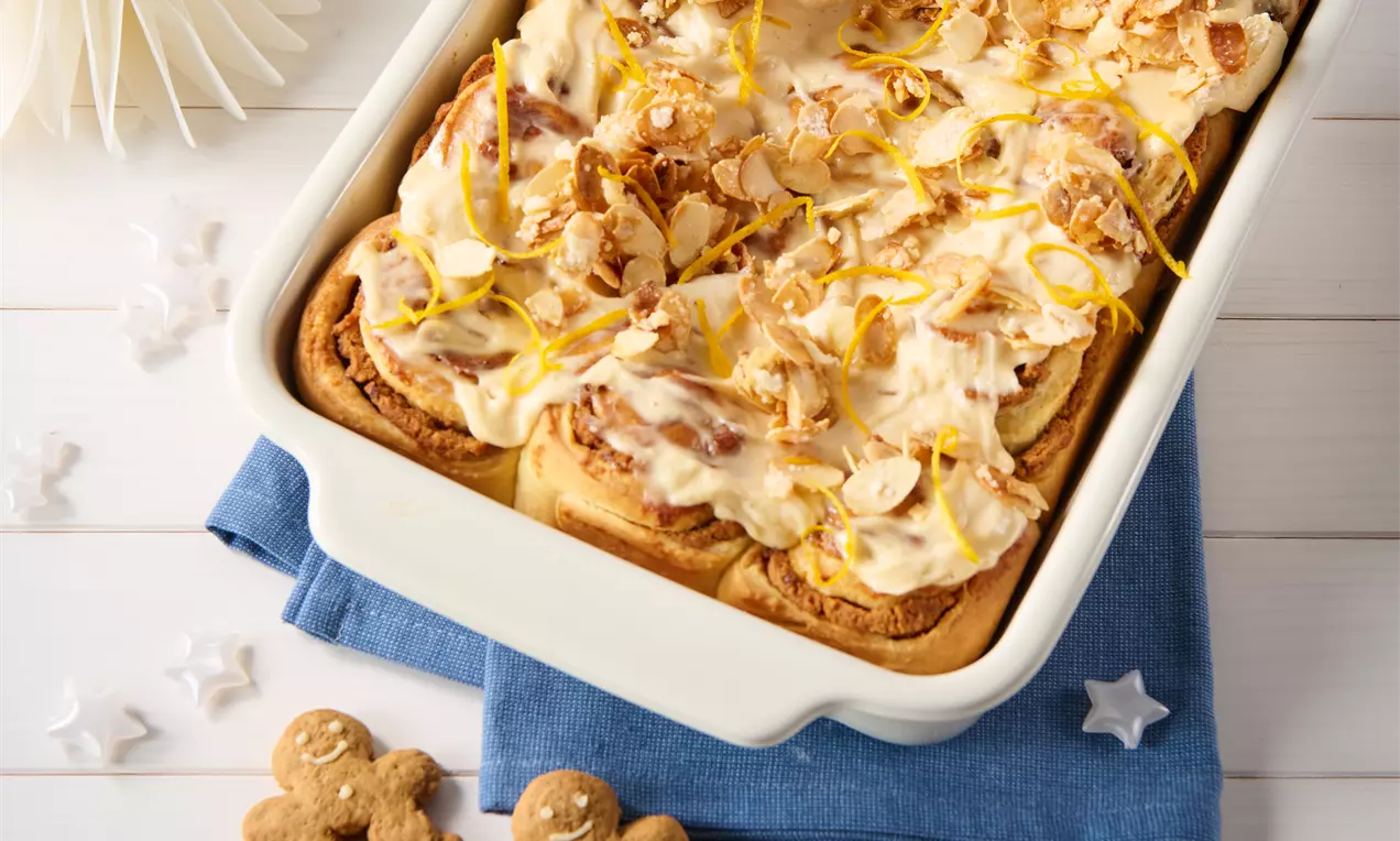 Saftige Lebkuchen-Zimtschnecken in einer weißen Form, bestrichen mit glatter Creme, garniert mit Mandelblättchen und frischen Zitronenzesten