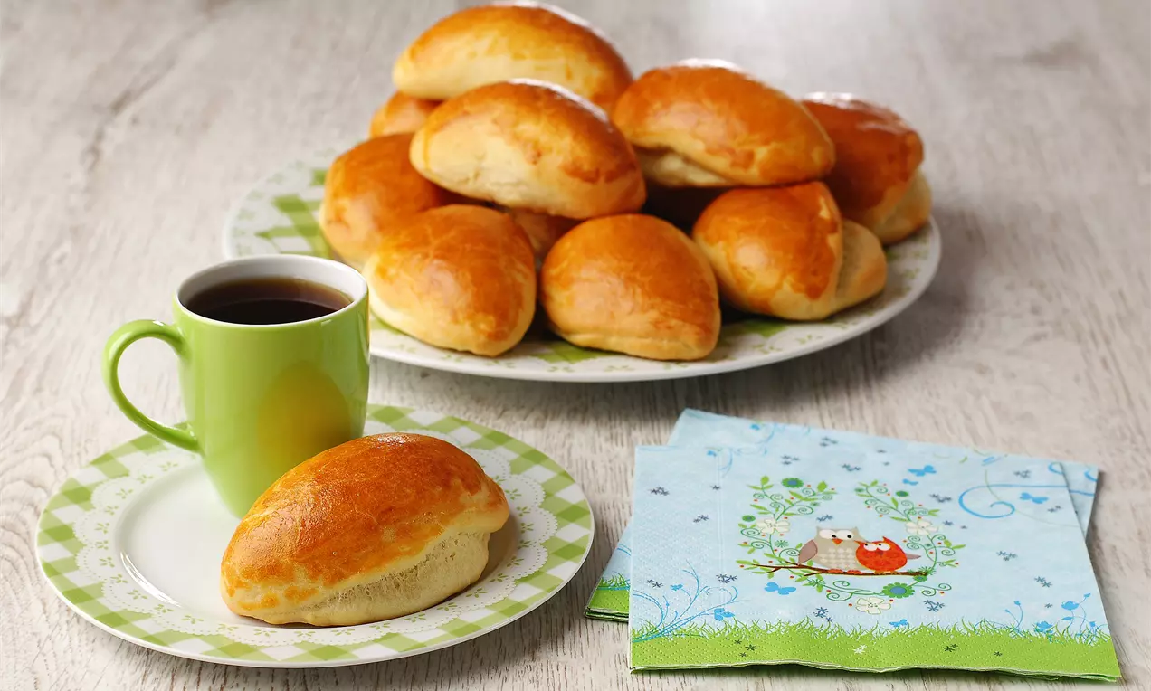 Goldgelb glänzende, halbmondförmige Pogaca-Schafkäse-Brötchen auf weißen Tellern, mit grünem Kaffebecher und bunt bedruckter Serviette arrangiert