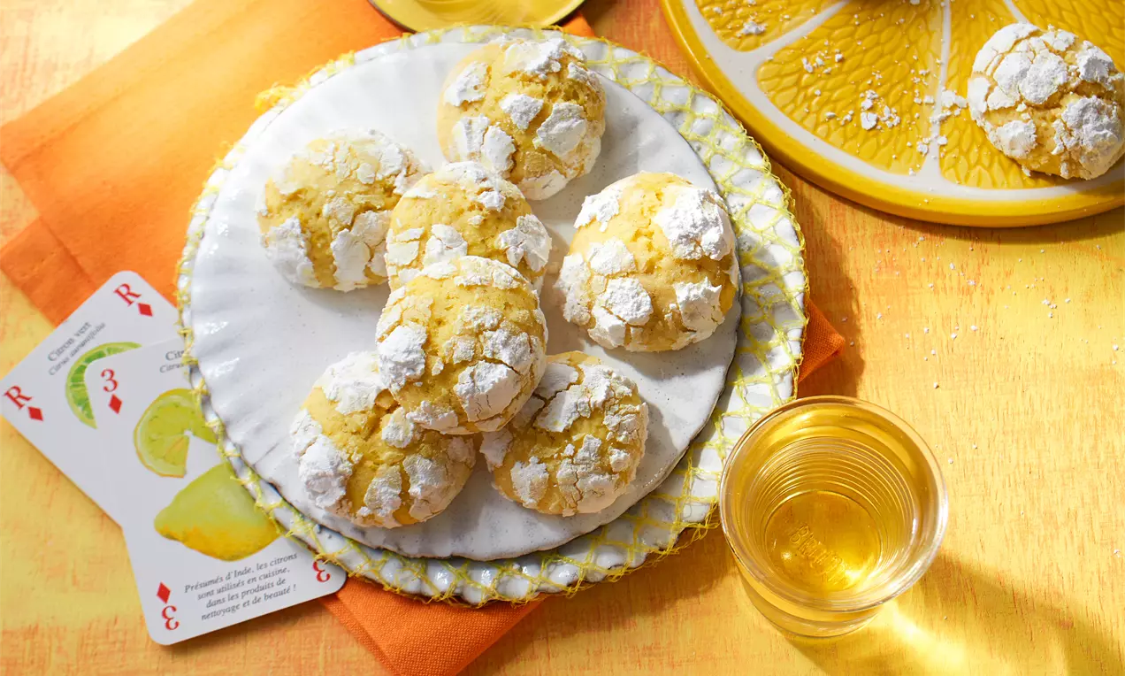 Zitronen-Crinkle-Cookies mit goldgelber, rissiger Oberfläche und weißem Puderzucker auf dekorativen Tellern neben Gläsern mit Getränk