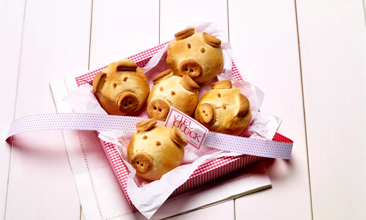 Goldbraune Schweinchenbrötchen mit runden Gesichtszügen und kleinen Teigohren in einer rot-weiß karierten Schale mit Papier ausgelegt
