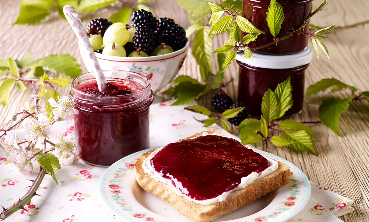 Samtige Brombeer-Stachelbeer-Konfitüre im Glas, tiefviolett glänzend, mit Brombeeren, Stachelbeeren und einer Zimtstange auf Holzbrett