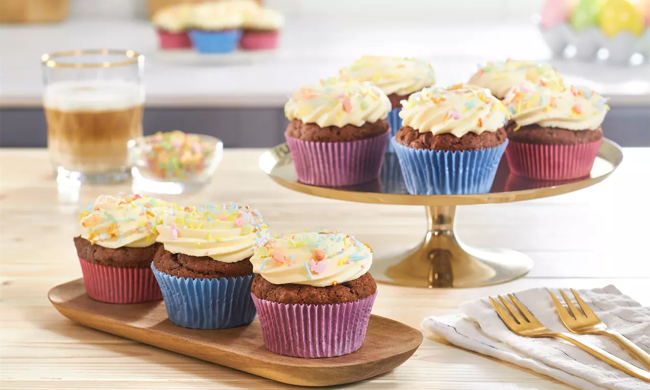Eierlikör-Cupcakes in farbigen Papierförmchen mit cremigem Topping und bunten Streuseln auf Holztablett und goldener Tortenplatte arrangiert