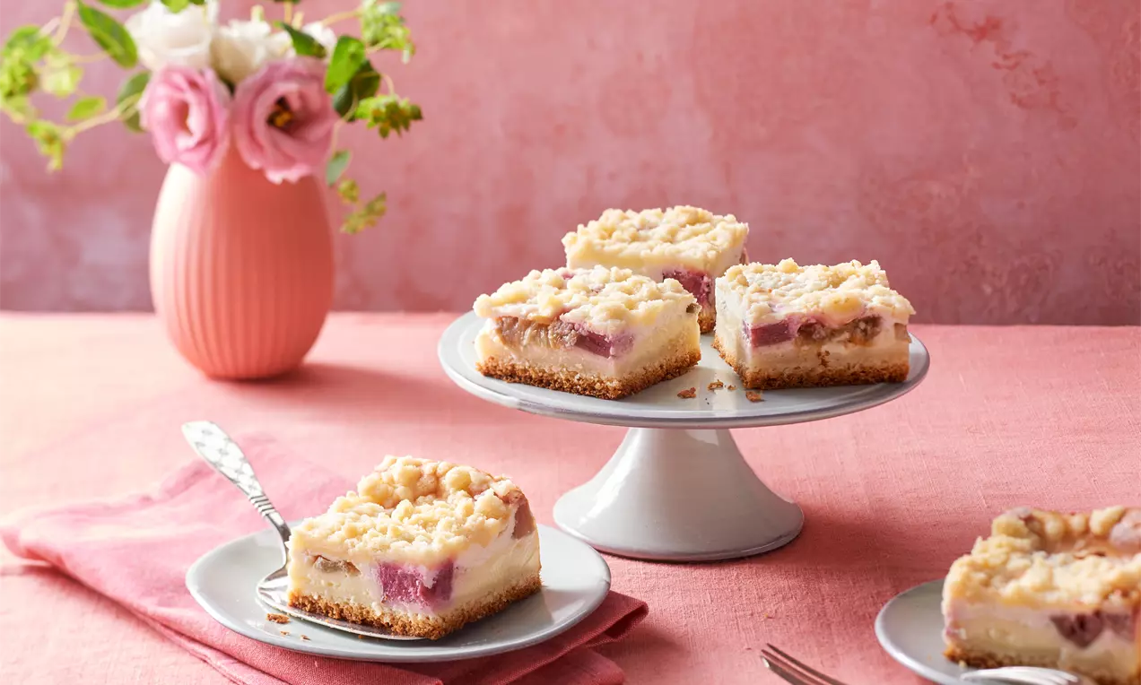 Rhabarber-Pudding-Käsekuchen mit goldenen Streuseln auf braunem Boden, serviert auf silbernen Tellern und einer weißen Tortenplatte