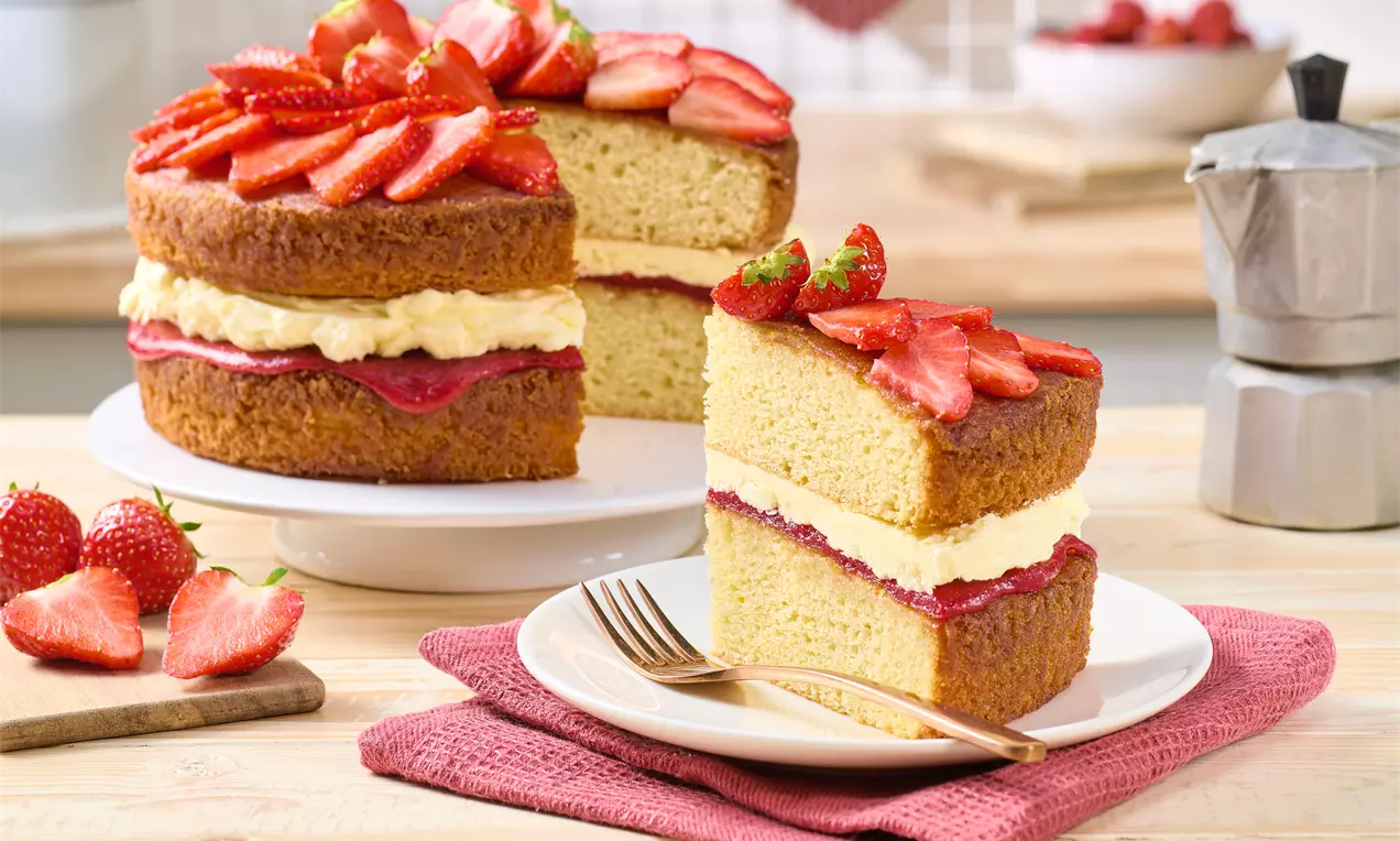 Victoria Sponge Cake auf einer weißen Platte, gefüllt mit Creme und Erdbeermarmelade, dekoriert mit frischen Erdbeerscheiben. Ein Stück liegt auf einem Teller mit Gabel, daneben weitere Erdbeeren.