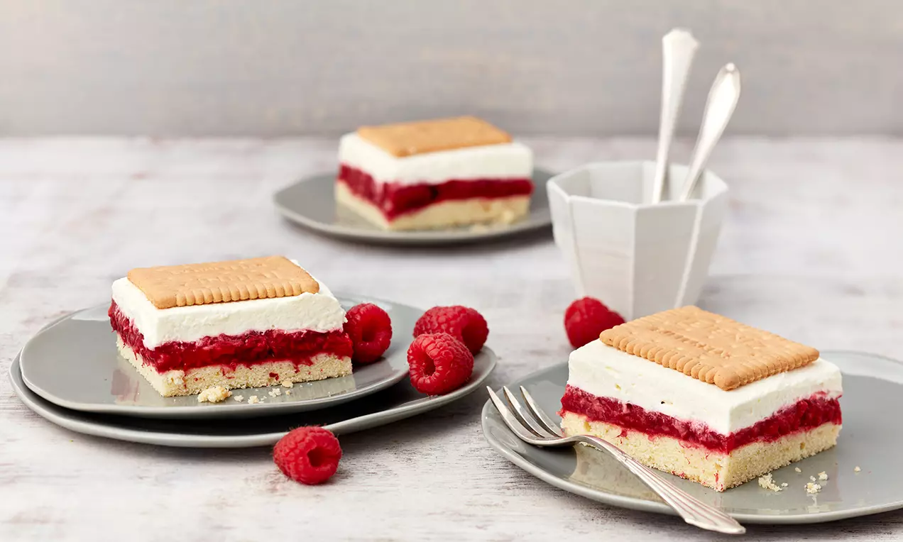 Rechteckige Butterkekskuchenstücke mit weißer Creme, roter Himbeerfüllung und lockerem Boden auf grauen Tellern, dekoriert mit frischen Himbeeren