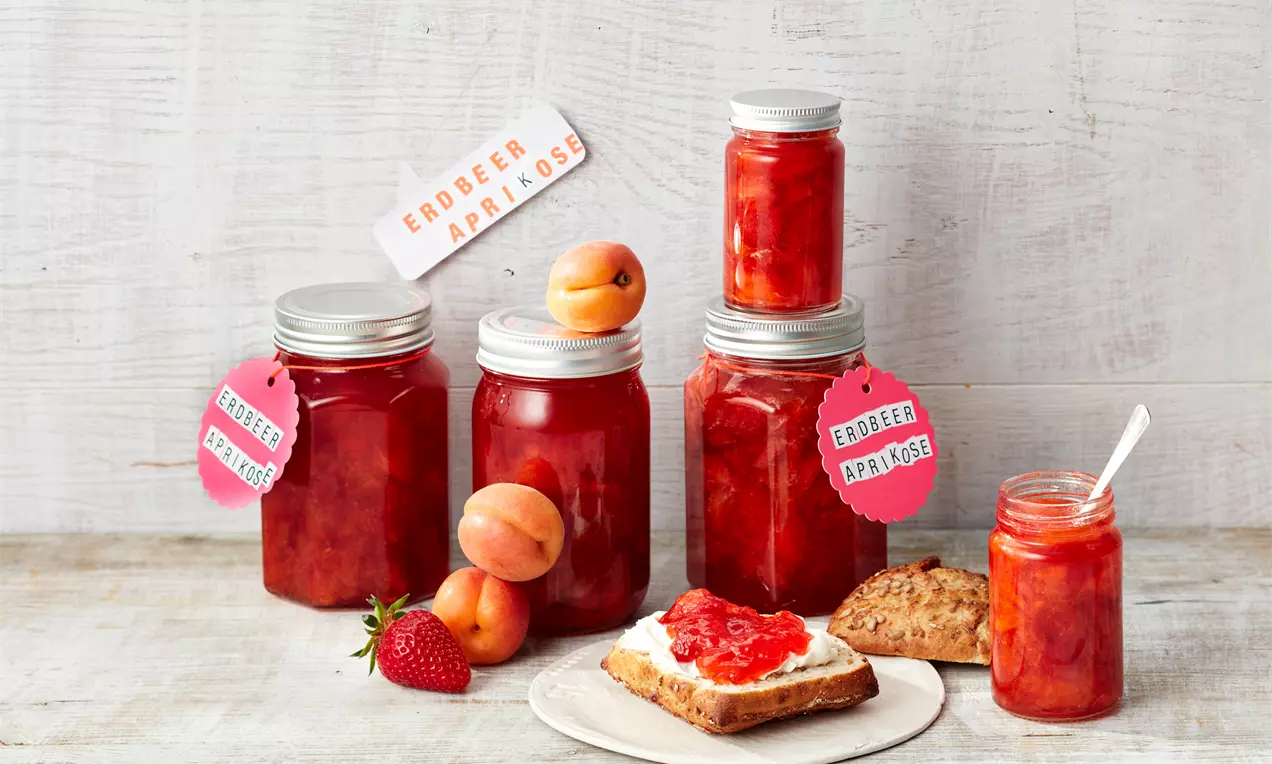 Gläser mit glänzend roter Erdbeer-Aprikosen-Konfitüre, beschrifteten Anhängern, frischen Früchten und angerichtetem Brot auf hellem Holztisch
