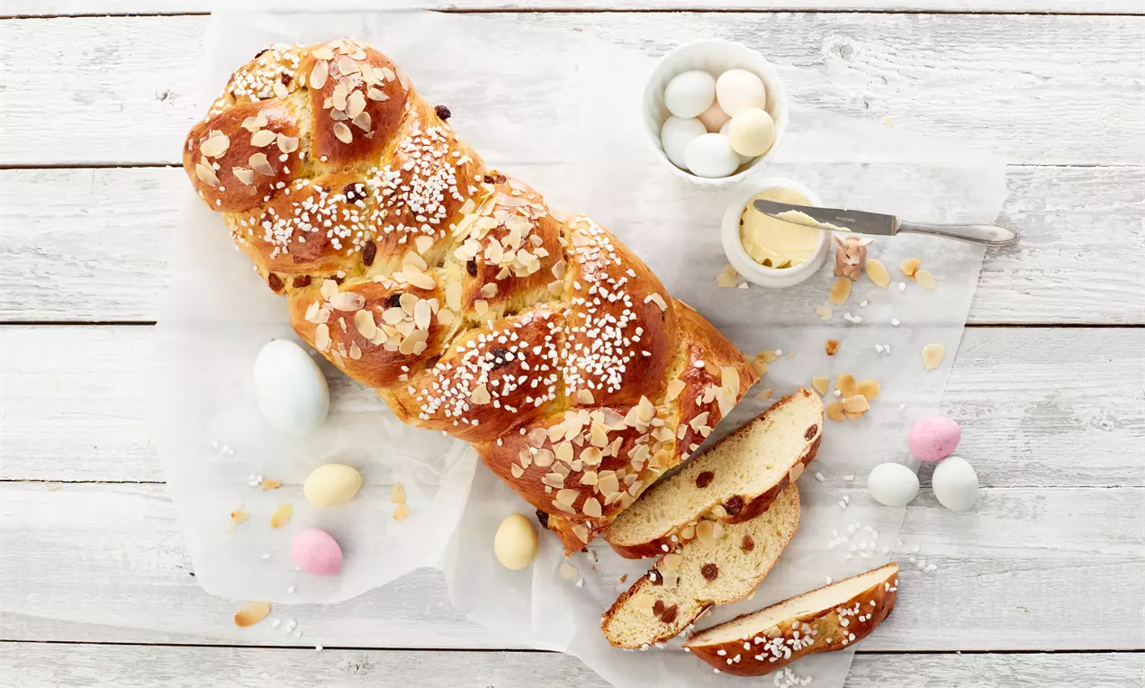 Goldbrauner Osterzopf mit Mandelsplittern und Hagelzucker auf Backpapier, daneben geschnittene Stücke, bunte Ostereier und eine Creme in Schale