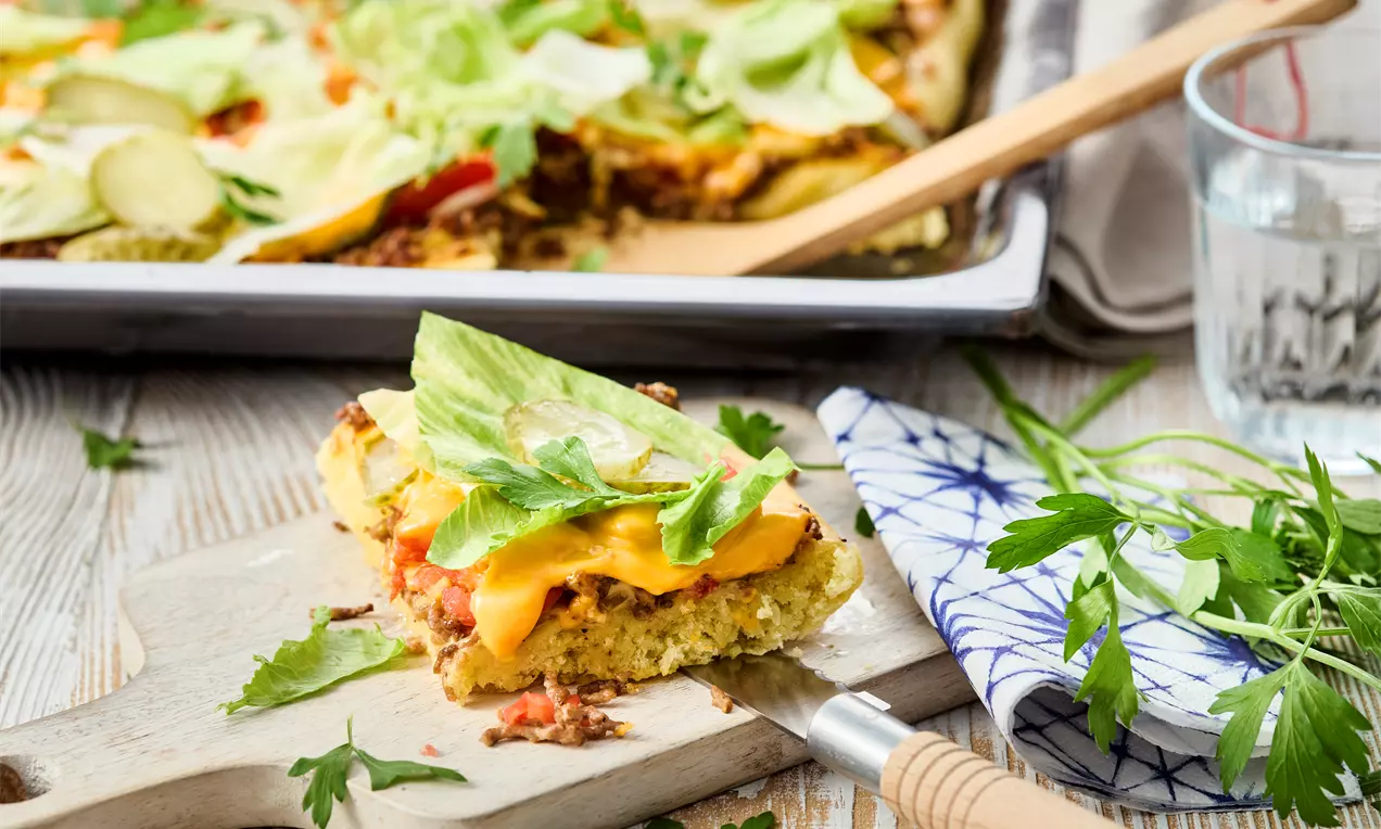 Eine goldbraune Ecke Cheeseburger-Pizza mit geschmolzenem Käse, Salat und Petersilie auf einem Holzbrett neben frischen Kräutern