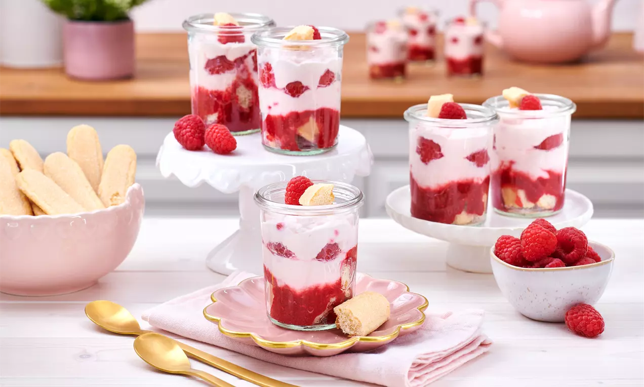 Schichtdessert aus weißer Mascarponecreme, roten Himbeeren und goldgelben Löffelbiskuits in Gläsern, dekoriert mit frischen Himbeeren