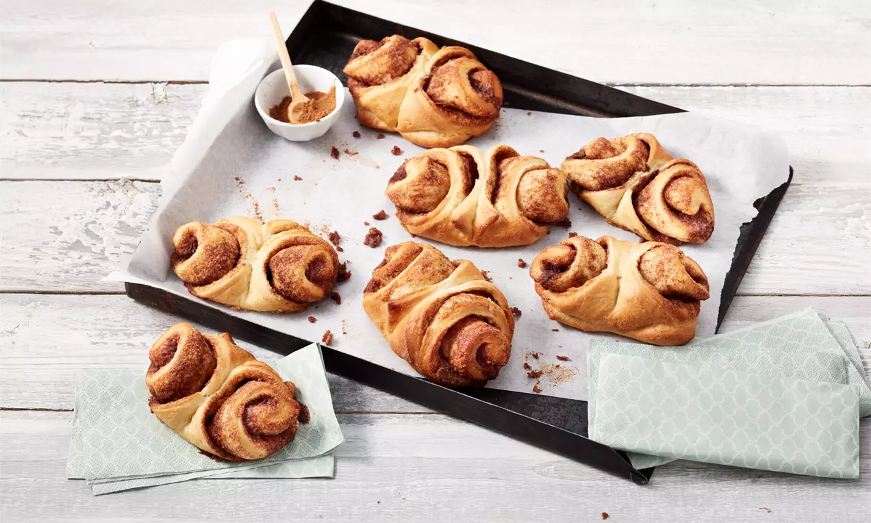 Goldbraune Franzbrötchen mit geschwungenen Zimtschichten auf Backpapier in schwarzem Blech, daneben Schale mit Zimtcreme und grüne Servietten
