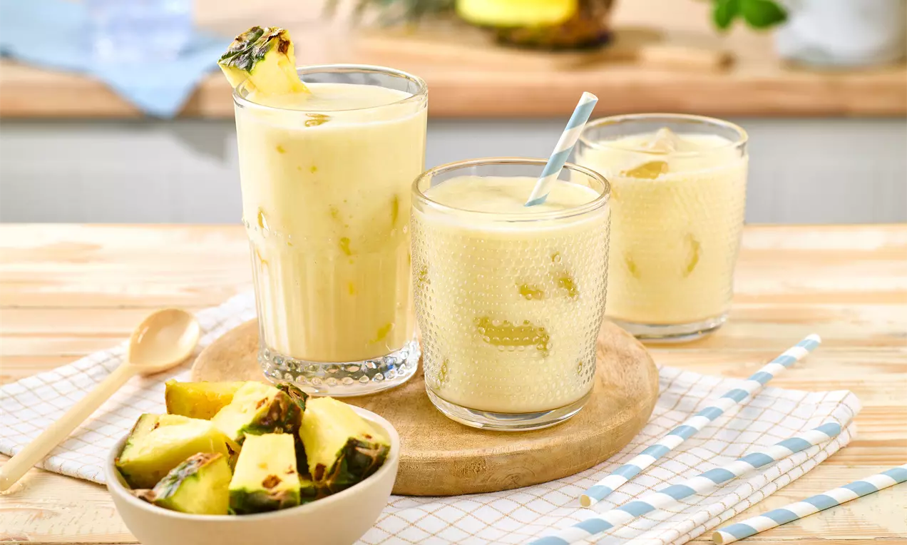 Cremiger Ananas-Shake in gläsernen Bechern, mit Stückchen und Strohhalm, serviert auf Holzbrett neben frischen Ananasstücken