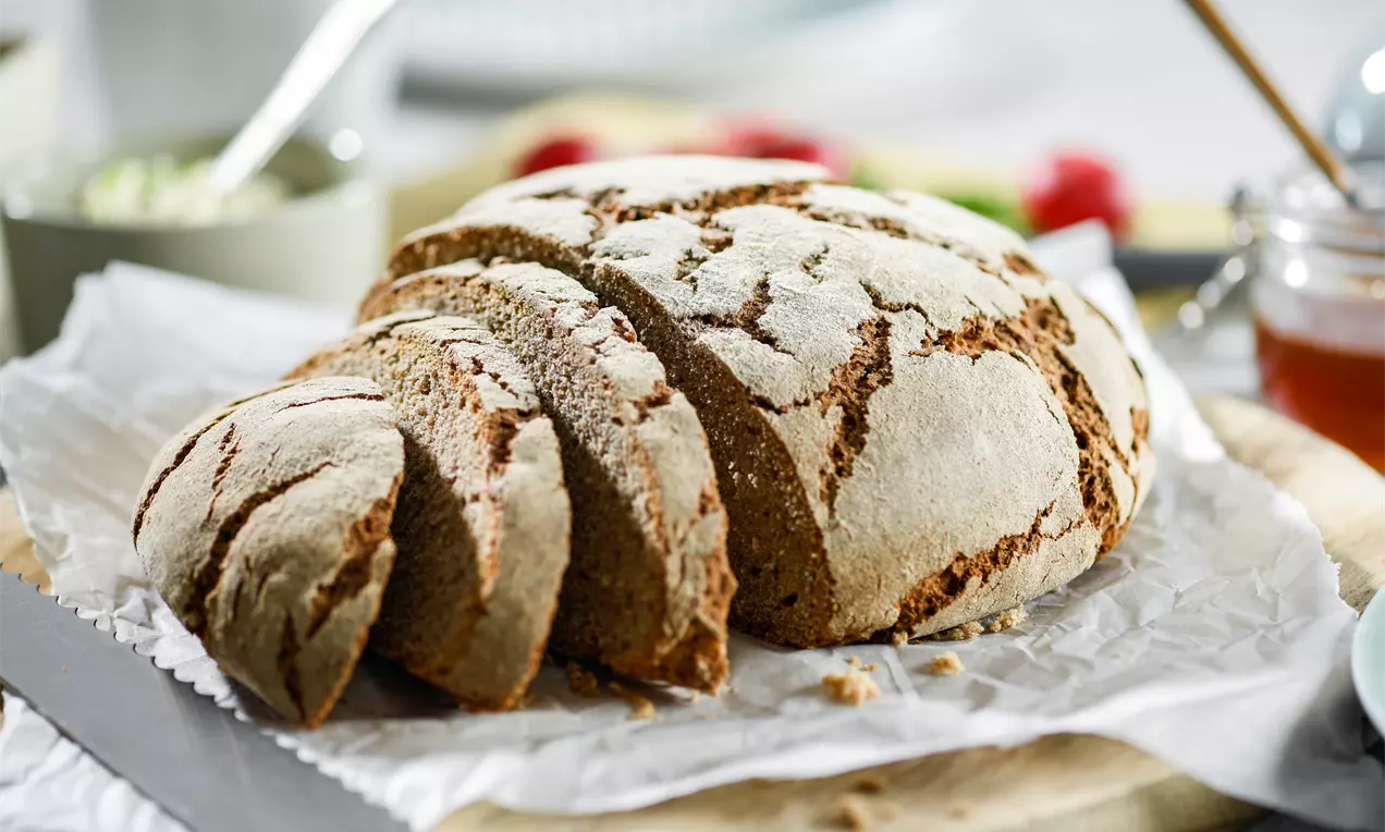 Roggenbrot mit grober Kruste, in dicke Scheiben geschnitten, auf Backpapier liegend, umgeben von Honigglas, Messer und Beilagen