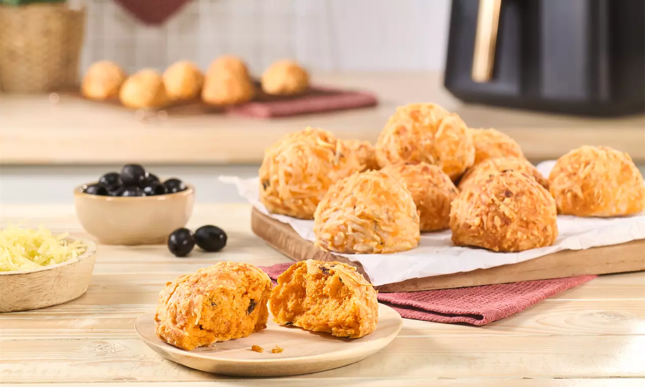 Mehrere goldbraune Pizzabrötchen aus dem Airfryer auf einem Holzbrett, im Vordergrund ein aufgebrochenes Brötchen auf einem Teller. Daneben Schalen mit geriebenem Käse und schwarzen Oliven.