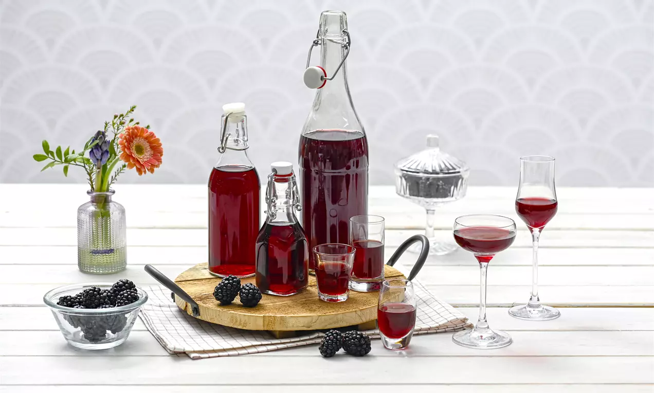 Tiefroter Brombeer-Likör in eleganten Glasflaschen und schmalen Gläsern auf einem Holzbrett, garniert mit frischen Brombeeren und Blumen