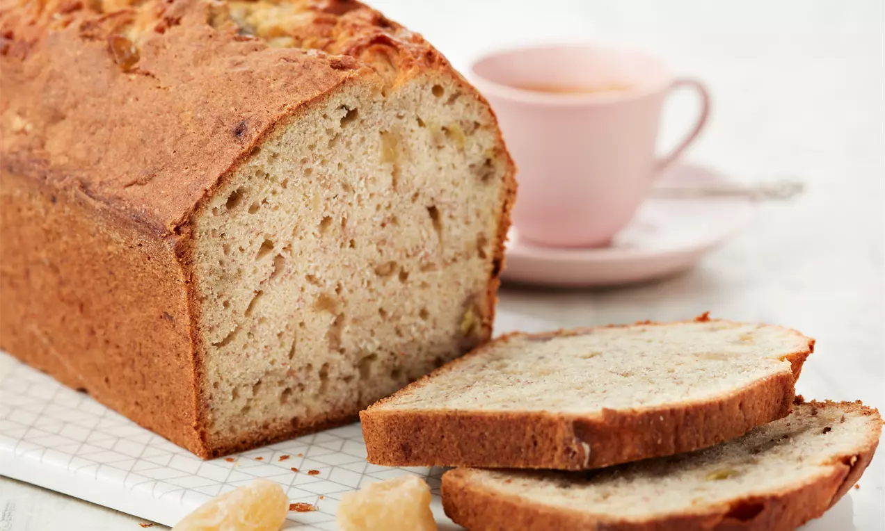Saftiges Bananenbrot mit goldbrauner Kruste, angeschnitten auf einem weißen Gitterbrett, neben zwei Scheiben und kandierten Ingwerstücken