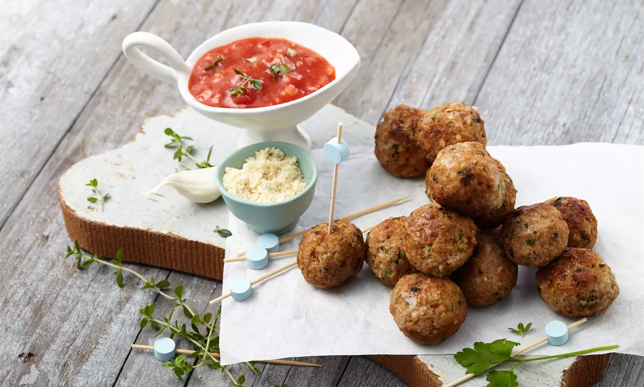 Hackbällchen mit goldbrauner Kruste auf Pergamentpapier serviert, daneben pikante Tomatensoße in weißer Sauciere und Parmesan in mintgrünem Schälchen