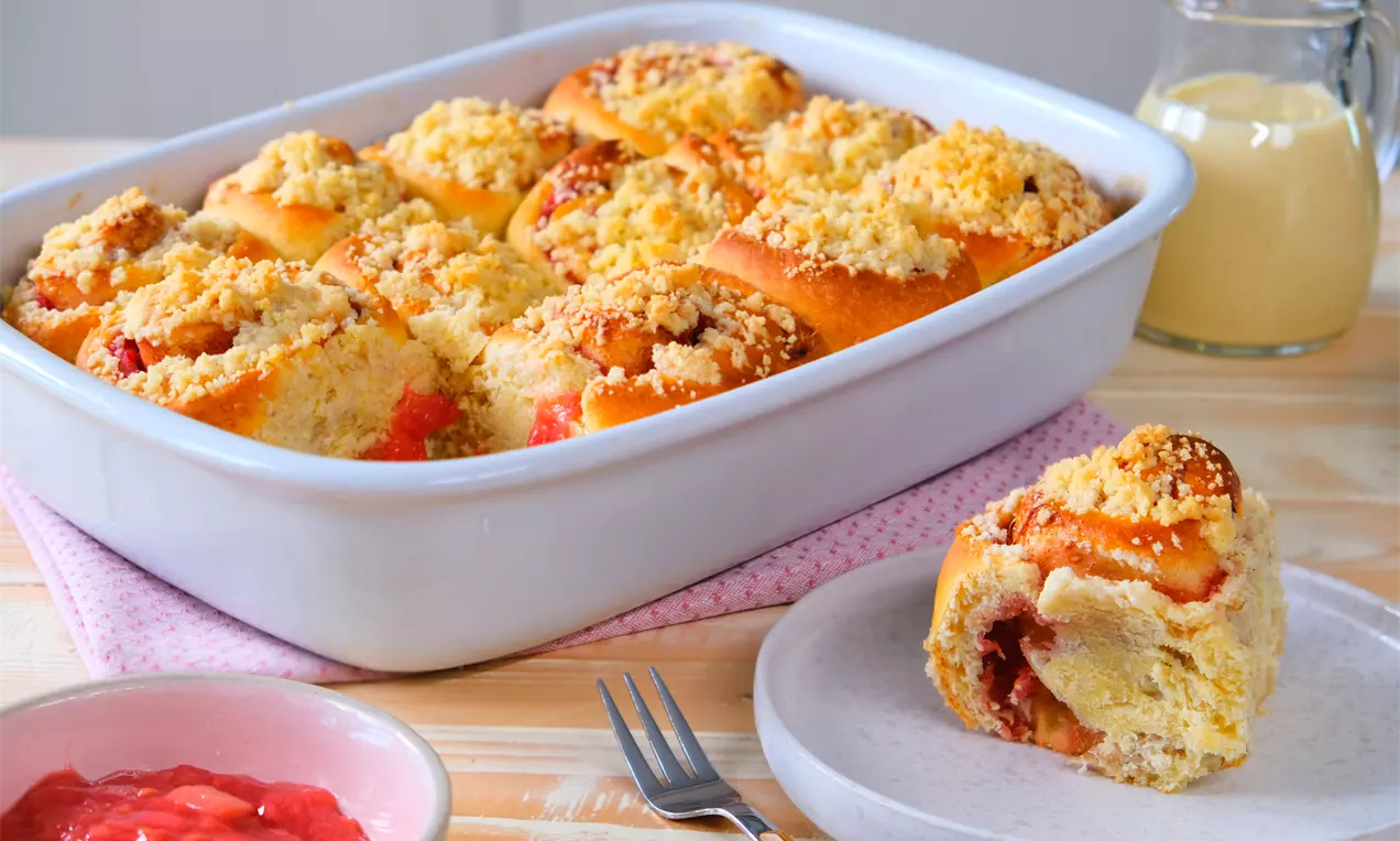 Goldbraune Rhabarber-Zimtschnecken mit Streuseln in einer weißen Auflaufform, daneben eine einzelne Schnecke auf einem Teller mit Kompott und Sauce